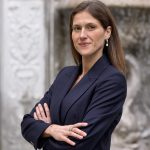 Una donna con lunghi capelli castani, che indossa un blazer blu navy e orecchini di perle, si trova all'esterno con le braccia incrociate e sorride con sicurezza. Sullo sfondo c'è una fontana o un muro decorativo in pietra.