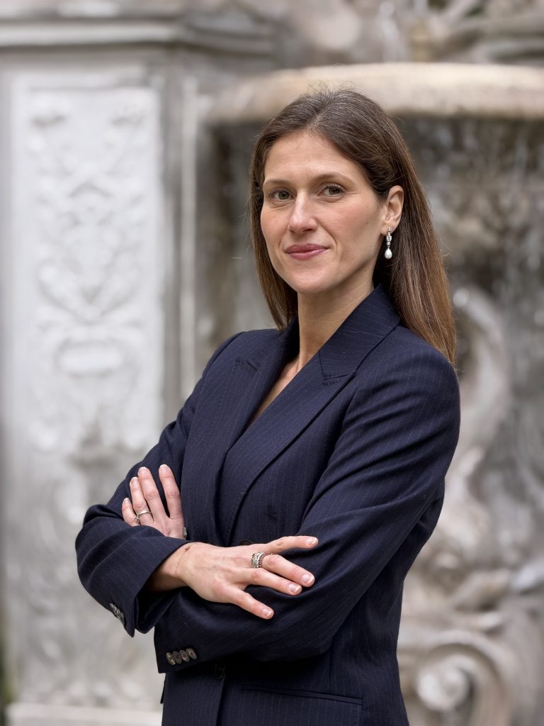 Una donna con lunghi capelli castani, che indossa un blazer blu navy e orecchini di perle, si trova all'esterno con le braccia incrociate e sorride con sicurezza. Sullo sfondo c'è una fontana o un muro decorativo in pietra.
