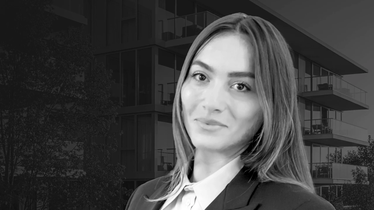 A woman with straight hair wearing a blazer and collared shirt smiles in front of a modern building. The image is in black and white.