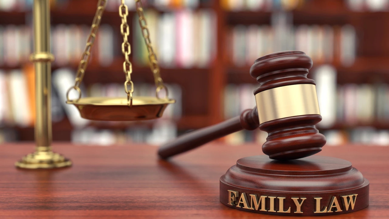 Un martelletto di legno con la scritta "DIRITTO DI FAMIGLIA" è appoggiato su una scrivania accanto a una bilancia di ottone, con file di scaffali sfocati sullo sfondo, a simboleggiare questioni legali come il diritto di famiglia e la convivenza di fatto.