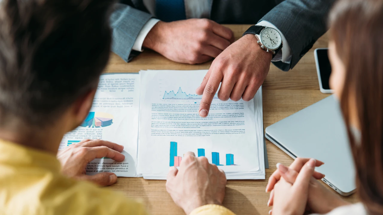 Tre persone siedono a un tavolo e discutono di documenti con grafici e tabelle. Una persona indica un rapporto stampato, mentre gli altri ascoltano attentamente, esaminando i documenti di un possibile family office.