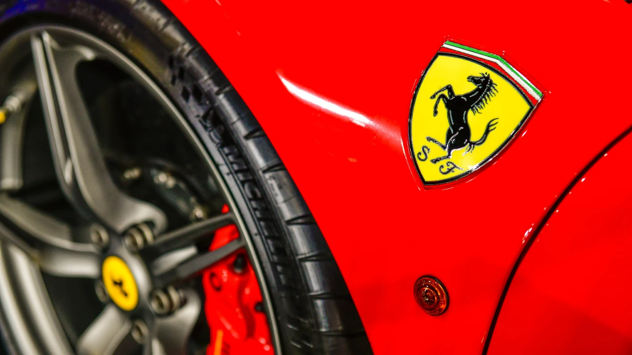 Primo piano di un'auto rossa Ferrari che mostra il logo giallo Ferrari con un cavallino rampante sul parafango, simbolo della fiducia della Ferrari, accanto a una ruota con un pneumatico nero visibile e parte del cerchio argentato. Il riferimento va a Ferrari, patti parasociali e trust: il controllo nel lungo periodo
