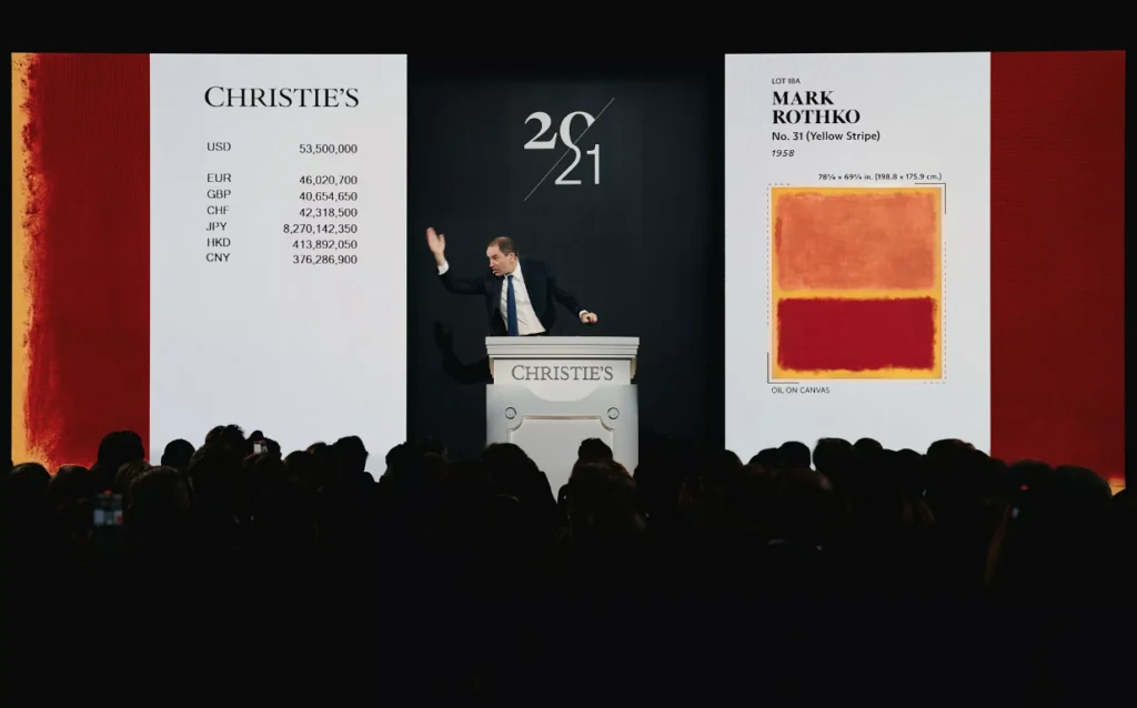 Un banditore d'asta è in piedi su un podio di Christie's, gesticolando verso un grande schermo che mostra il dipinto di Mark Rothko "No. 7 (Yellow Stripe)", con i prezzi delle offerte visualizzati in varie valute davanti a un pubblico seduto.