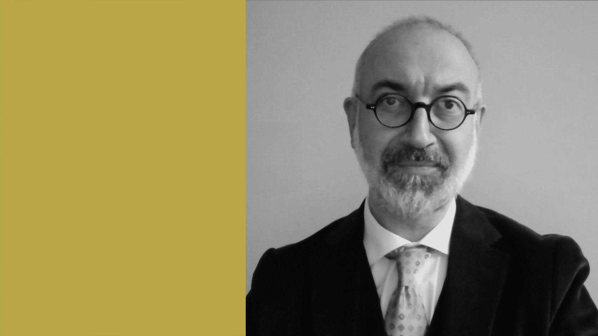Un uomo con occhiali, barba e baffi, in giacca e cravatta, posa su uno sfondo semplice. Sul lato sinistro dell'immagine è presente un grande rettangolo giallo.
