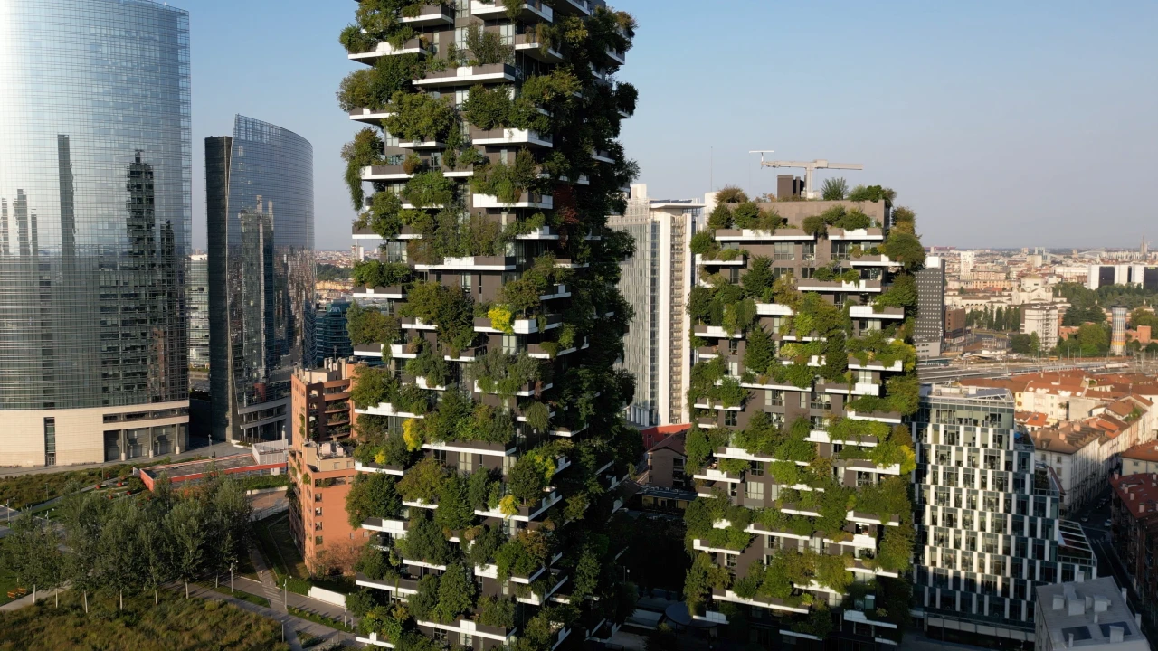 Two modern high-rise buildings covered with lush green plants and trees on balconies, surrounded by other skyscrapers and city buildings under a clear sky.