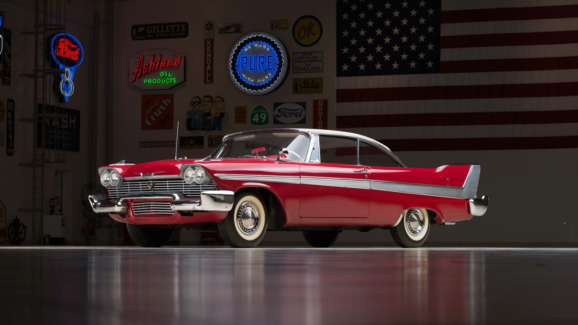 Un'auto d'epoca rossa e bianca è parcheggiata all'interno su un pavimento lucido, con insegne retrò e una grande bandiera americana sulla parete dietro di essa.