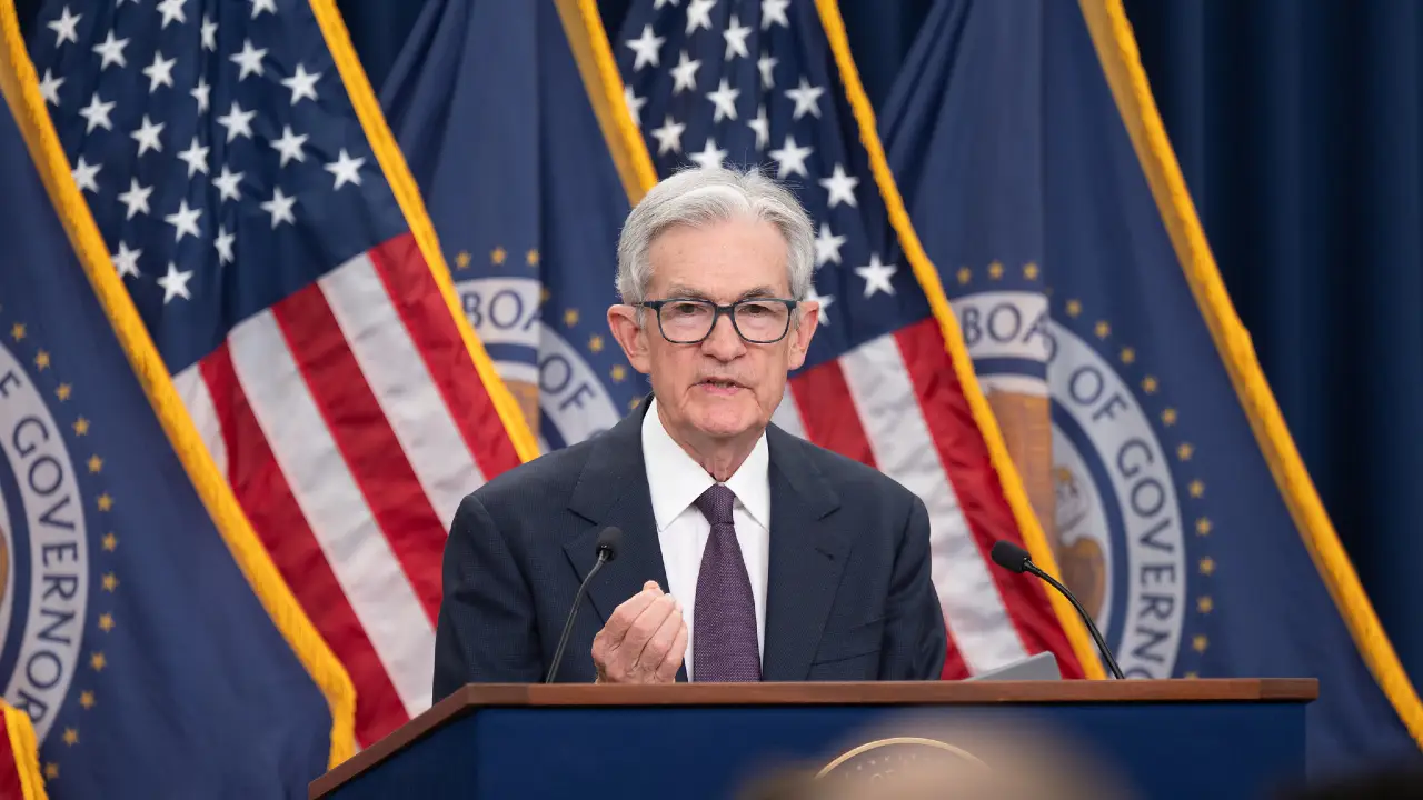 Un uomo in giacca e cravatta e occhiali parla su un podio davanti a diverse bandiere degli Stati Uniti e a striscioni della Federal Reserve.