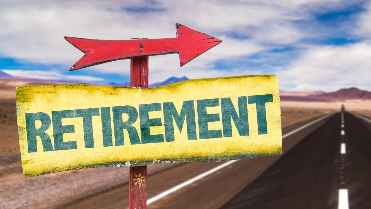 A red arrow sign with a yellow board reading "RETIREMENT" points the way along an empty road in a desert landscape, symbolizing the journey toward retirement beneath a blue sky with clouds.