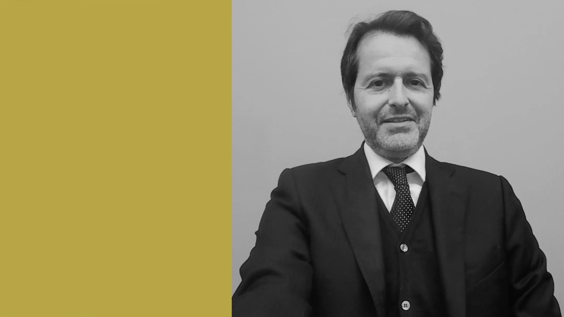 Un uomo in giacca e cravatta, con capelli scuri e barba corta, siede su uno sfondo semplice. Il lato sinistro dell'immagine presenta un blocco di colore giallo senape. L'immagine è in bianco e nero, tranne il blocco giallo.