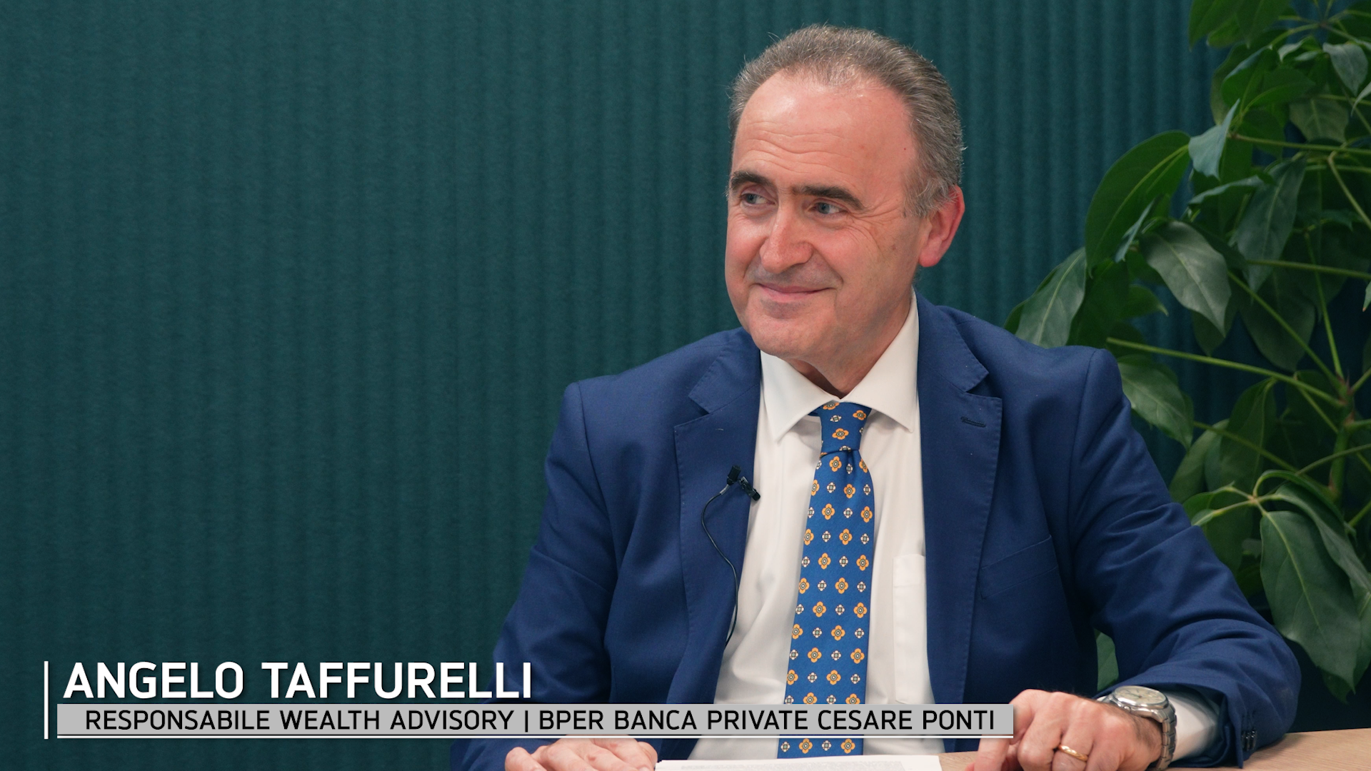 Un uomo in abito blu e cravatta a fantasia siede a un tavolo, sorridendo, con le mani appoggiate davanti a sé. Sullo sfondo c'è una pianta e una didascalia che recita "Angelo Taffurelli, Responsabile Wealth Advisory, BPER Banca Private Cesare Ponti".