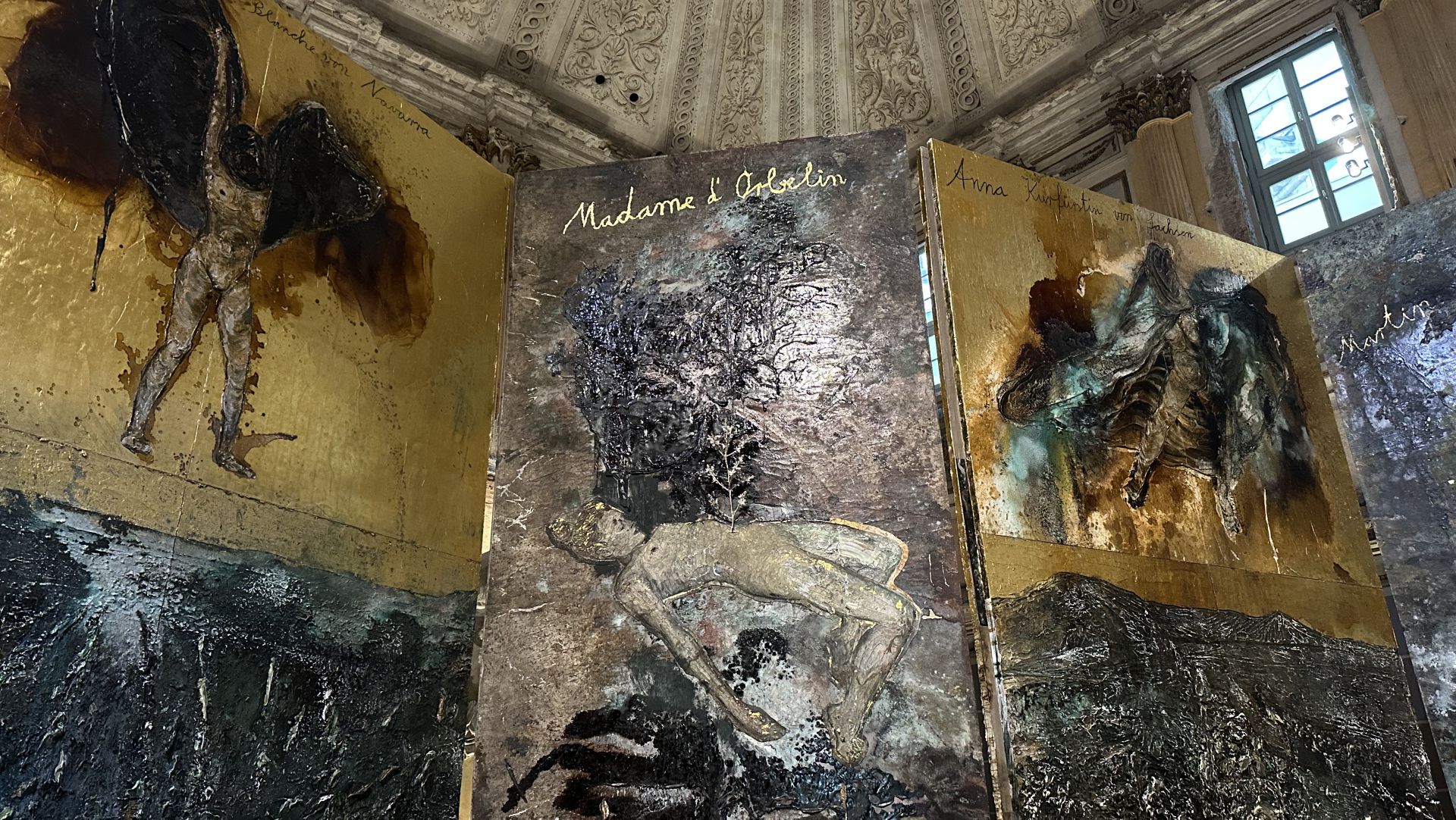Tre grandi dipinti con sfondi dorati e figure umane astratte e angeliche, che ricordano lo stile di Anselm Kiefer, sono esposti in fila all'interno di una sala ornata e dai soffitti alti. Il pannello centrale è intitolato "Madame l'Oiselin".