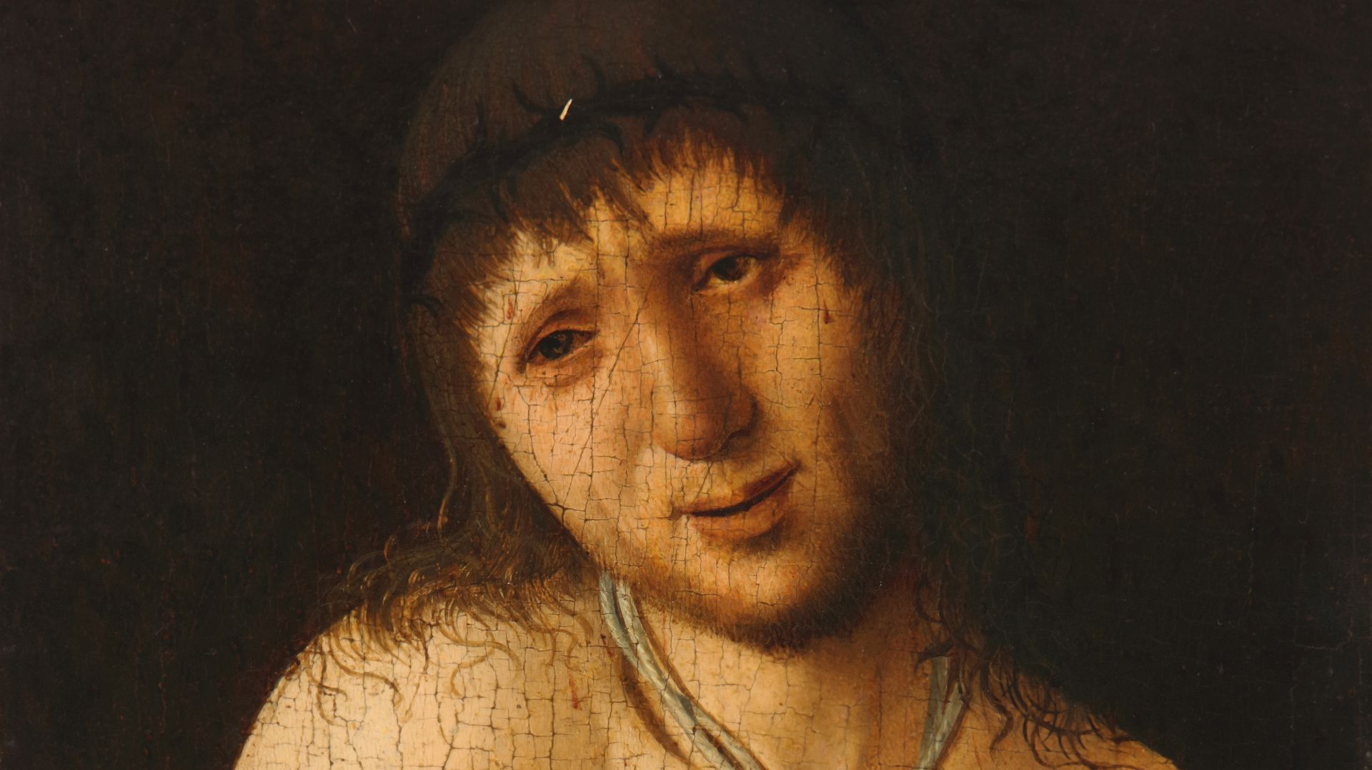 Un primo piano di un dipinto rinascimentale raffigura un uomo addolorato con una corona di spine, lunghi capelli castani e una superficie invecchiata e screpolata. I suoi occhi sono abbassati e la sua espressione è triste su uno sfondo scuro.