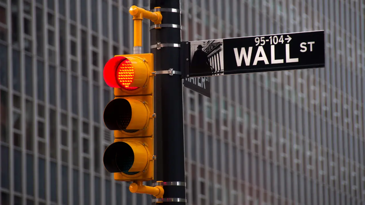 Un semaforo rosso è appeso accanto a un cartello stradale di Wall Street, con un alto edificio di uffici sullo sfondo.