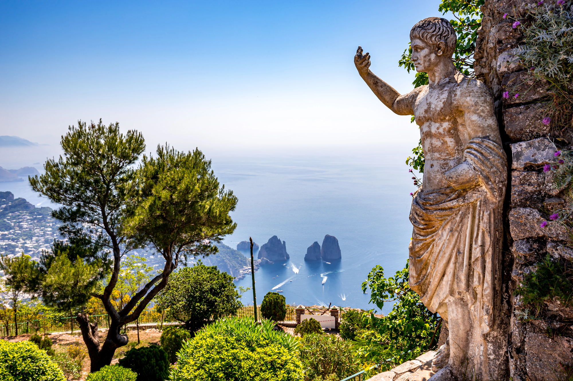 A weathered ancient statue stands on a hilltop, overlooking lush greenery and the blue sea with rock formations off the coast of Capri, Italy, under a clear sky.
