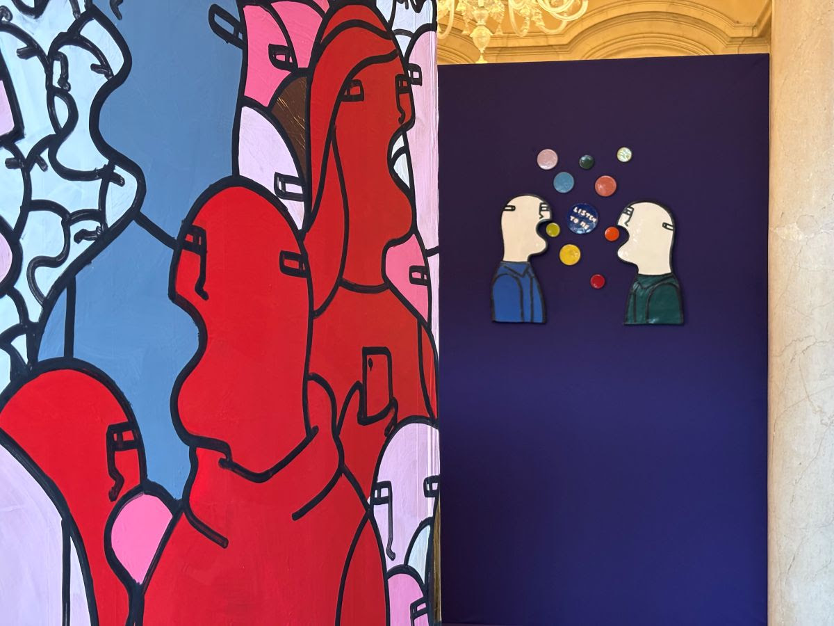 Una mostra d'arte colorata presenta figure astratte e audaci. A sinistra, volti dai contorni rossi e rosa si mescolano. A destra, due personaggi dal volto bianco si fronteggiano su una parete blu scuro con bolle vocali colorate sopra di loro.