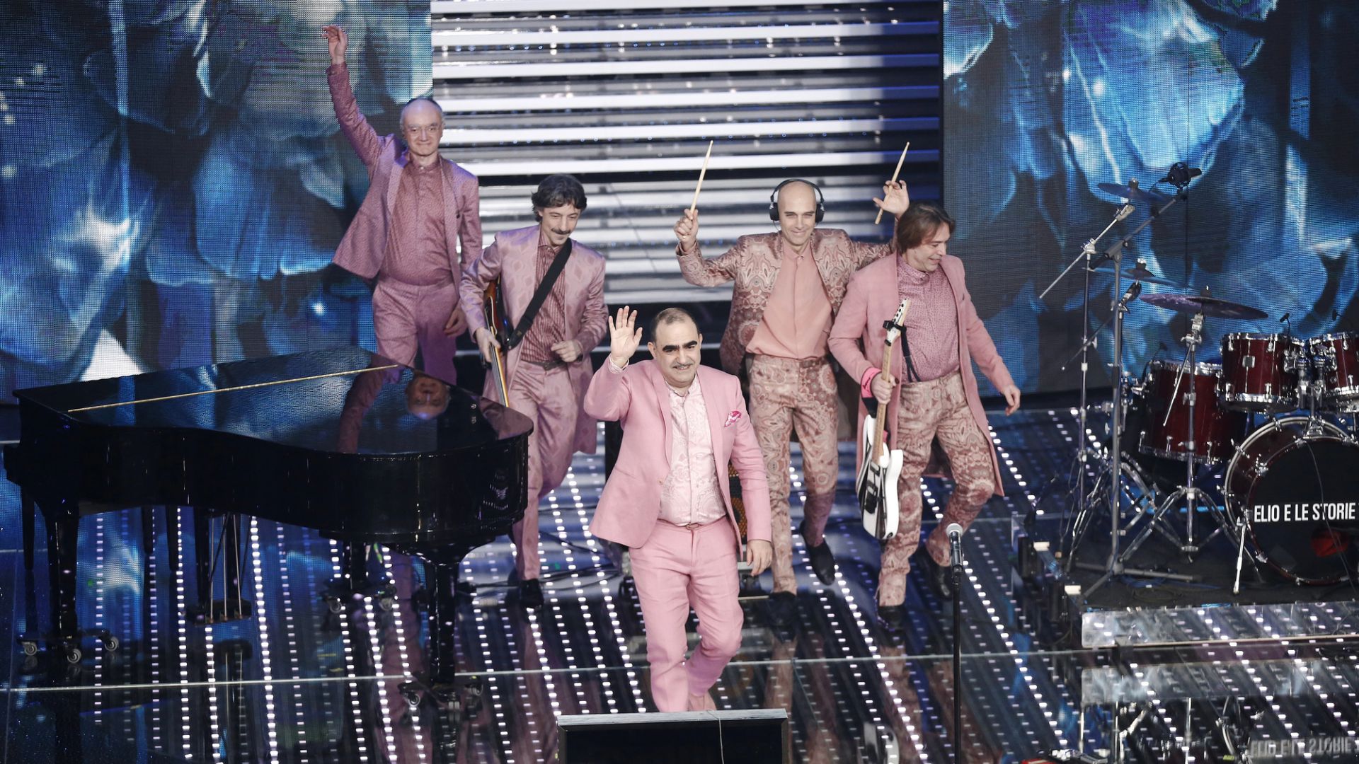 Una band di sei uomini vestiti con abiti rosa coordinati cammina su un palco, salutando e sorridendo, circondata da strumenti musicali tra cui un pianoforte a coda e una batteria. Il palcoscenico è illuminato da luci brillanti e da un fondale a motivi blu.