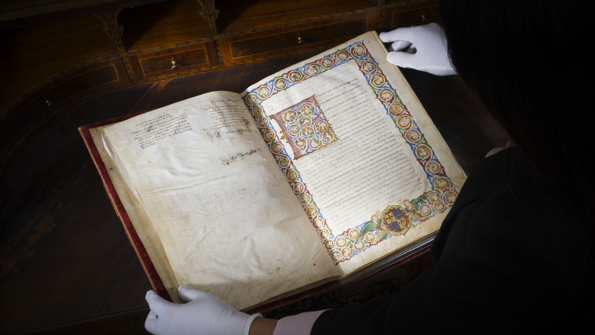 Una persona che indossa guanti bianchi apre con cura un manoscritto miniato con bordi colorati e intricati, esposto su un tavolo di legno.