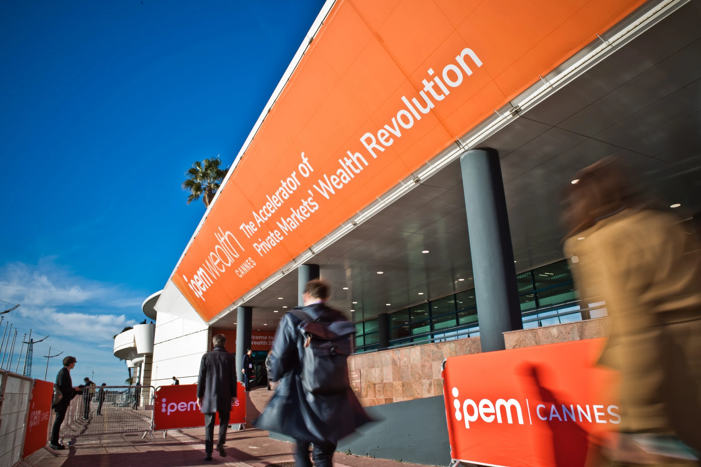 Le persone passano davanti a un edificio moderno con un grande striscione arancione che recita "The Accelerator of Private Markets' Wealth Revolution" e "iPEM Cannes" sotto un cielo azzurro e limpido.