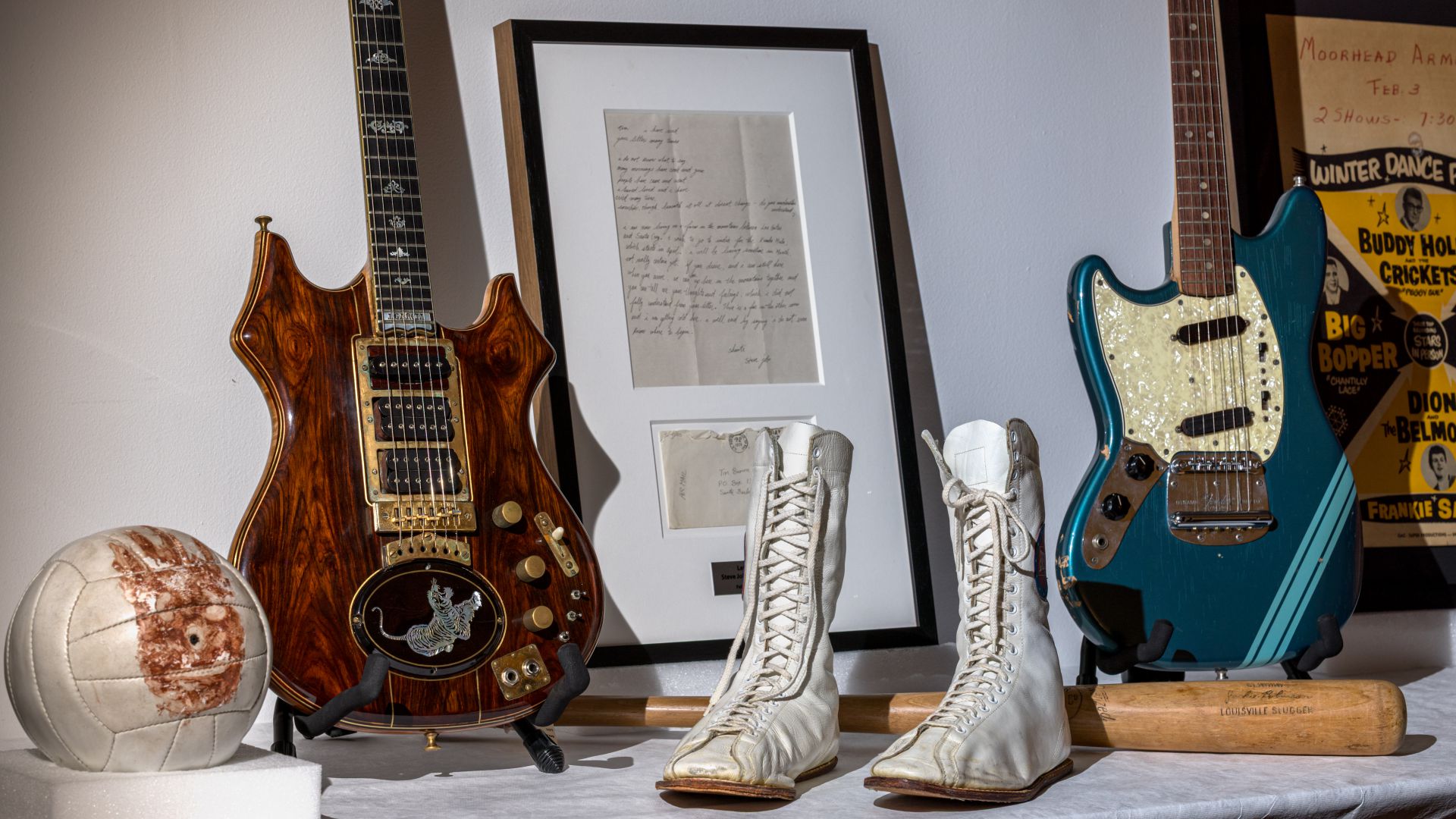 Un'esposizione con un pallone da calcio vintage, una chitarra elettrica marrone, lettere scritte a mano incorniciate, stivali bianchi con lacci, una chitarra elettrica blu e parte di un poster musicale sullo sfondo.