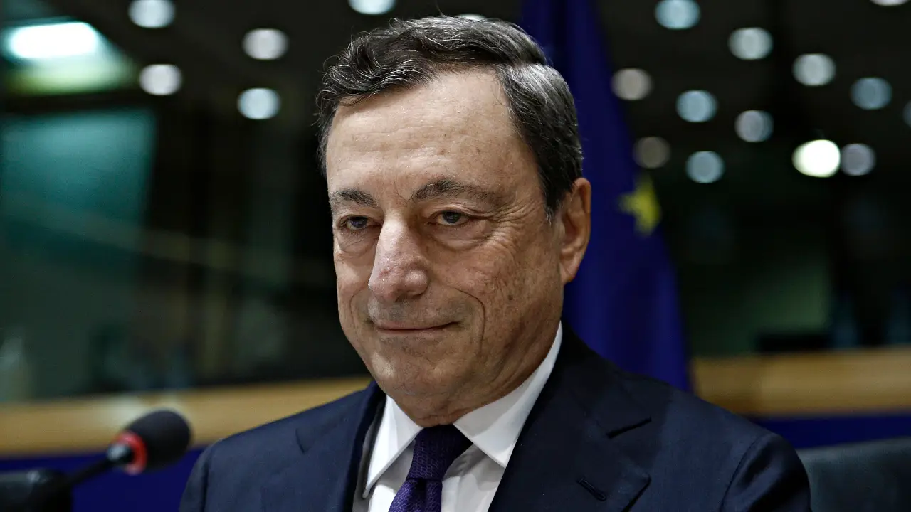 Un uomo in abito scuro e cravatta si trova davanti a uno sfondo sfocato con la bandiera dell'Unione Europea e le luci, guardando leggermente di lato con un'espressione neutra, che evoca la presenza composta di Mario Draghi mentre supervisiona le questioni del patrimonio.