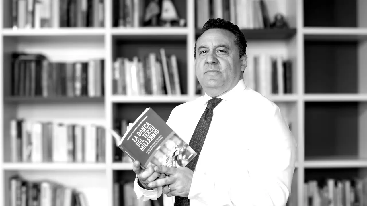 Un uomo in camicia bianca è in piedi davanti a una libreria, con in mano un libro aperto intitolato "La Banca del Tercer Milenio". L'immagine è in bianco e nero.