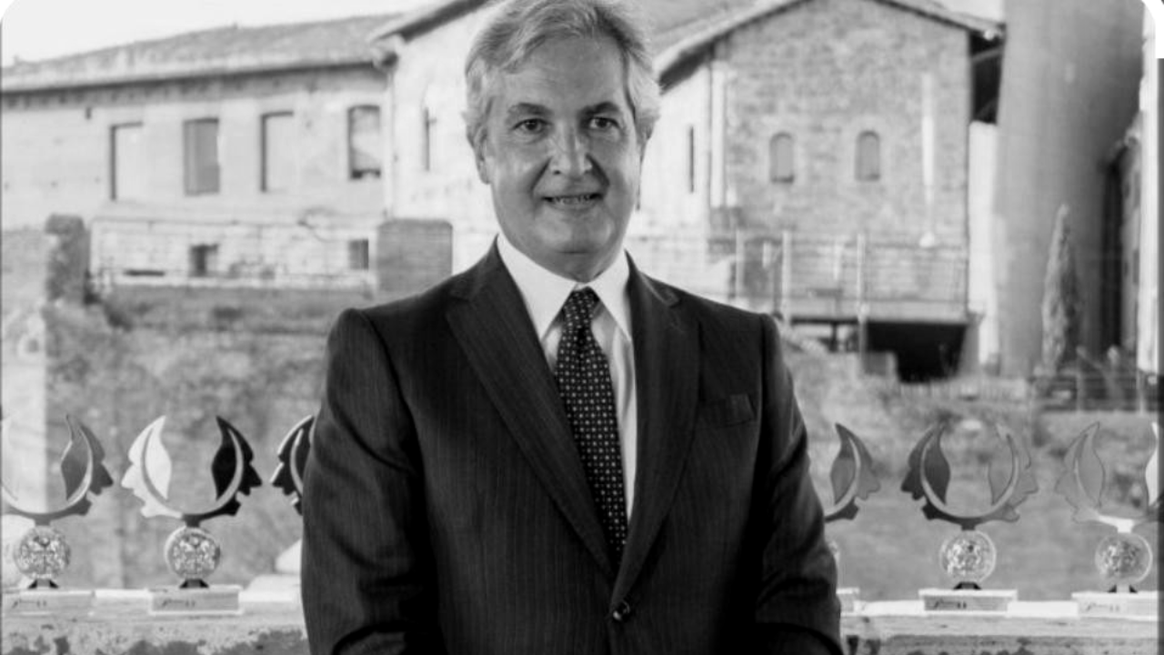 Un uomo in giacca e cravatta si trova davanti a una fila di trofei, con edifici storici in pietra e il logo della Pilloni Banca del Fucino visibili nell'immagine in bianco e nero.