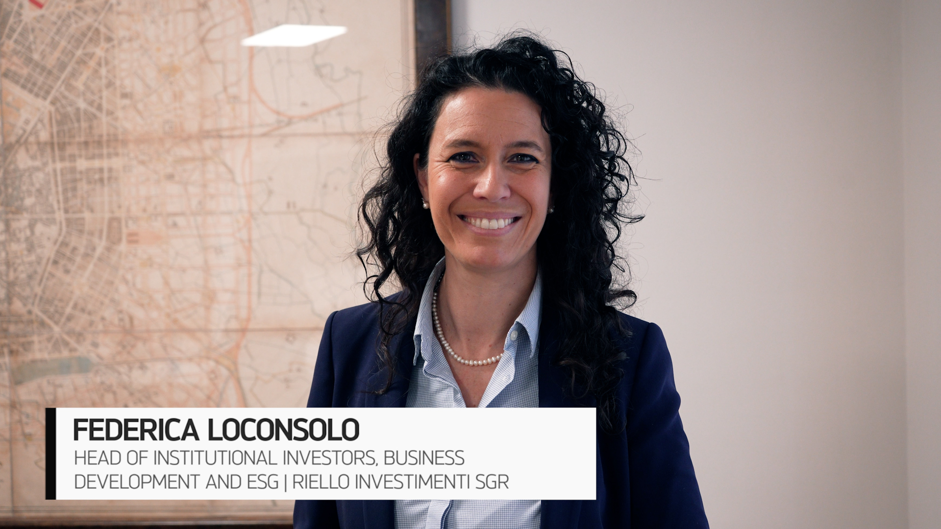 Federica Loconsolo di Riello Investimenti indossa un blazer blu e una collana di perle, sorride mentre si trova in casa. Una mappa è appesa alla parete dietro di lei.