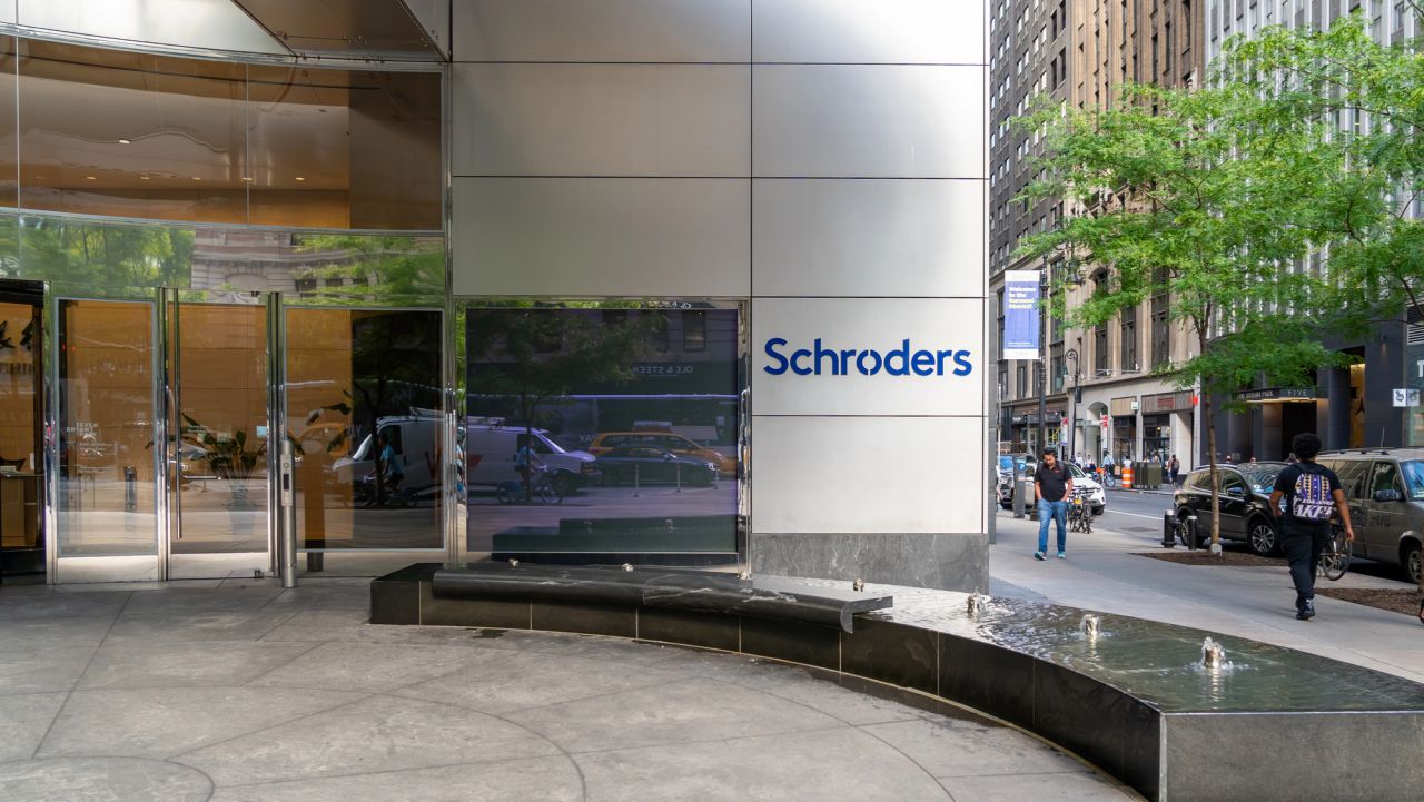 L'ingresso di un edificio moderno con l'insegna "Schroders" su una parete, con porte in vetro, una fontana d'acqua curva e una strada cittadina con auto e pedoni sullo sfondo.
