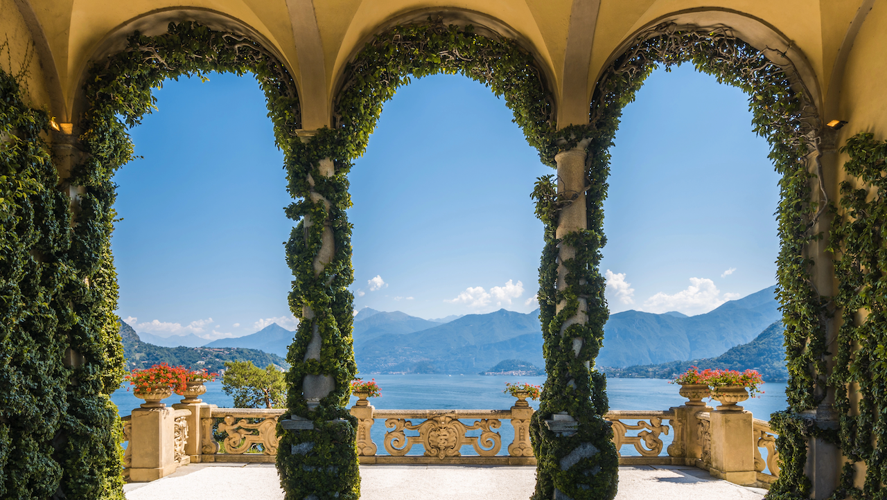Three ivy-covered stone arches frame a scenic view of a blue lake and distant mountains under a clear sky; flower pots with red blooms sit on the balustrade.