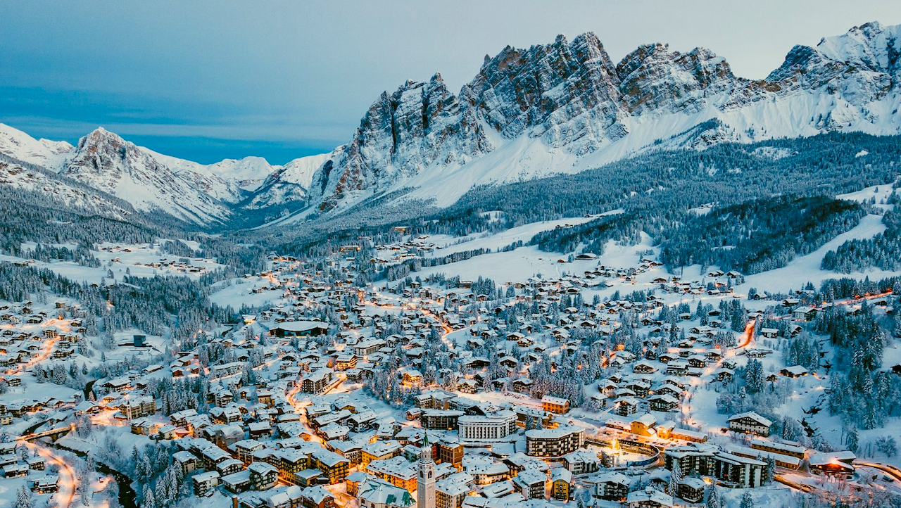 Una città alpina innevata, adagiata in una valle sotto alte e aspre montagne, al tramonto, con edifici e strade illuminati a giorno: uno scenario perfetto per un accogliente weekend a Cortina, circondato da fitti boschi e pendii innevati.