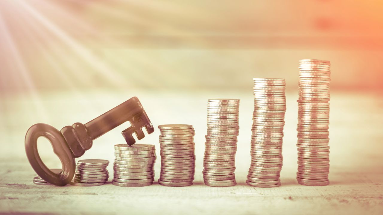 Una chiave d'epoca poggia su una piccola pila di monete, con accanto quattro pile di monete più alte disposte in ordine crescente, a simboleggiare la crescita finanziaria e il successo. La luce del sole entra dall'angolo in alto a sinistra.