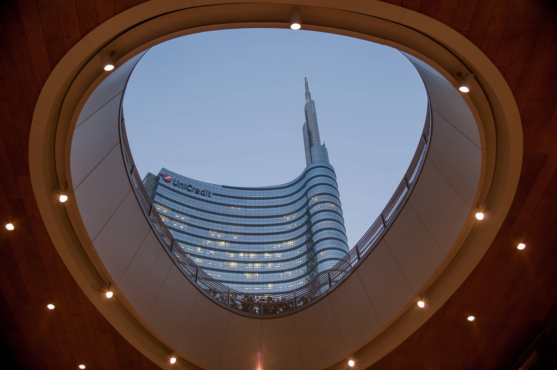 Vista di un moderno grattacielo di vetro attraverso un'apertura ovale nel soffitto, con luci calde intorno all'apertura e un cielo sereno sullo sfondo.