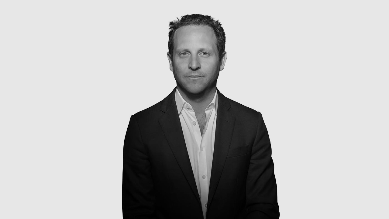 Teddy Mouawad stands against a plain, light gray background, facing the camera with a neutral expression. The image is in black and white.
