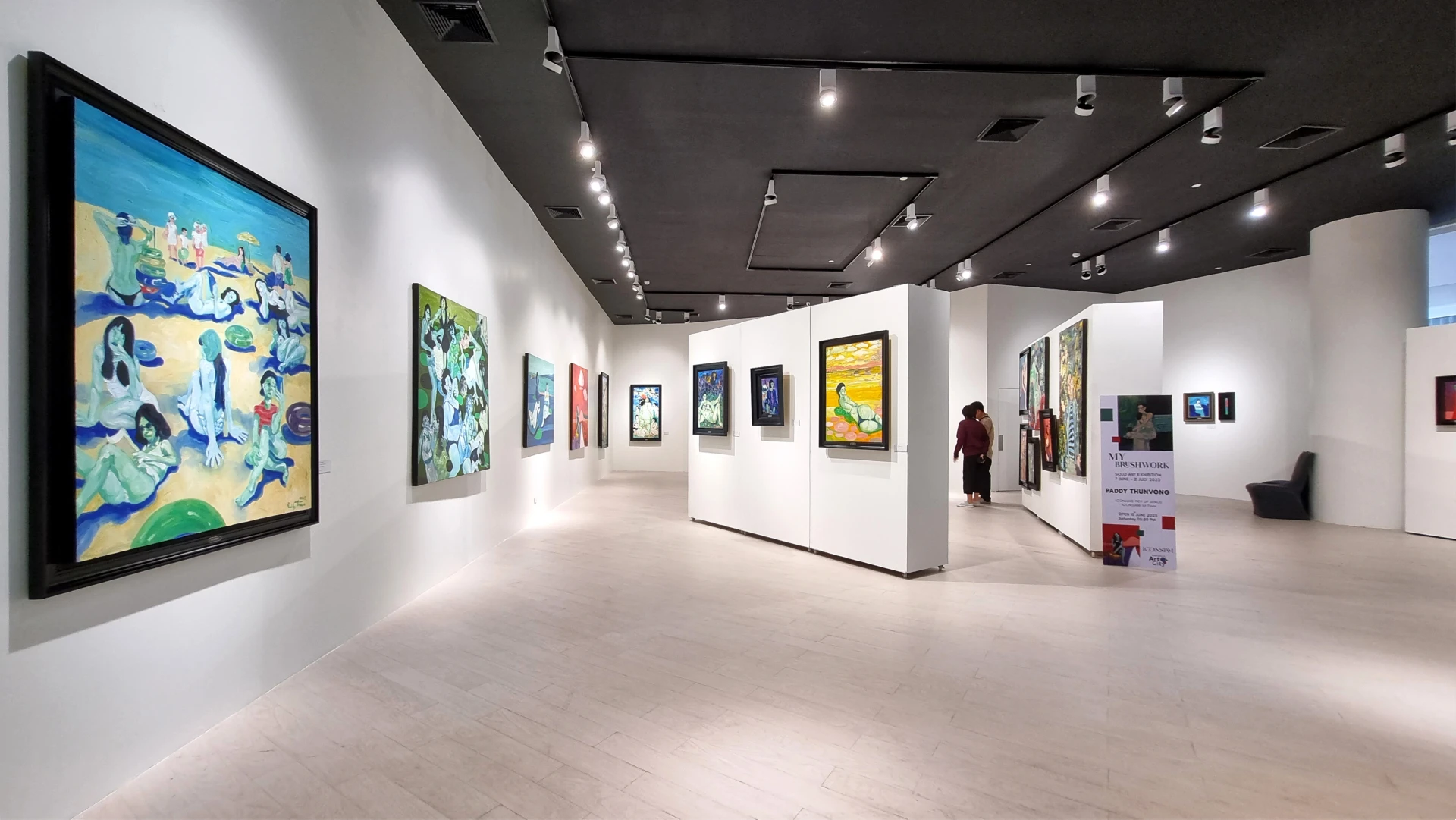 Luminosa galleria d'arte moderna con pareti bianche e pavimenti in legno chiaro, che espone dipinti colorati. Una persona si trova in lontananza vicino a un angolo con un cartello espositivo: uno spazio stimolante per investire nell'arte sotto le eleganti luci nere del soffitto.