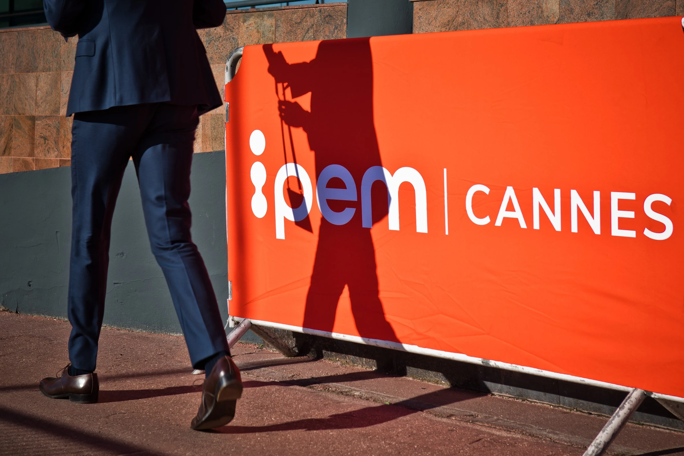 A person in a dark suit walks past an orange barrier with the white text "IPEM | CANNES"; their shadow and briefcase are visible on the barrier.