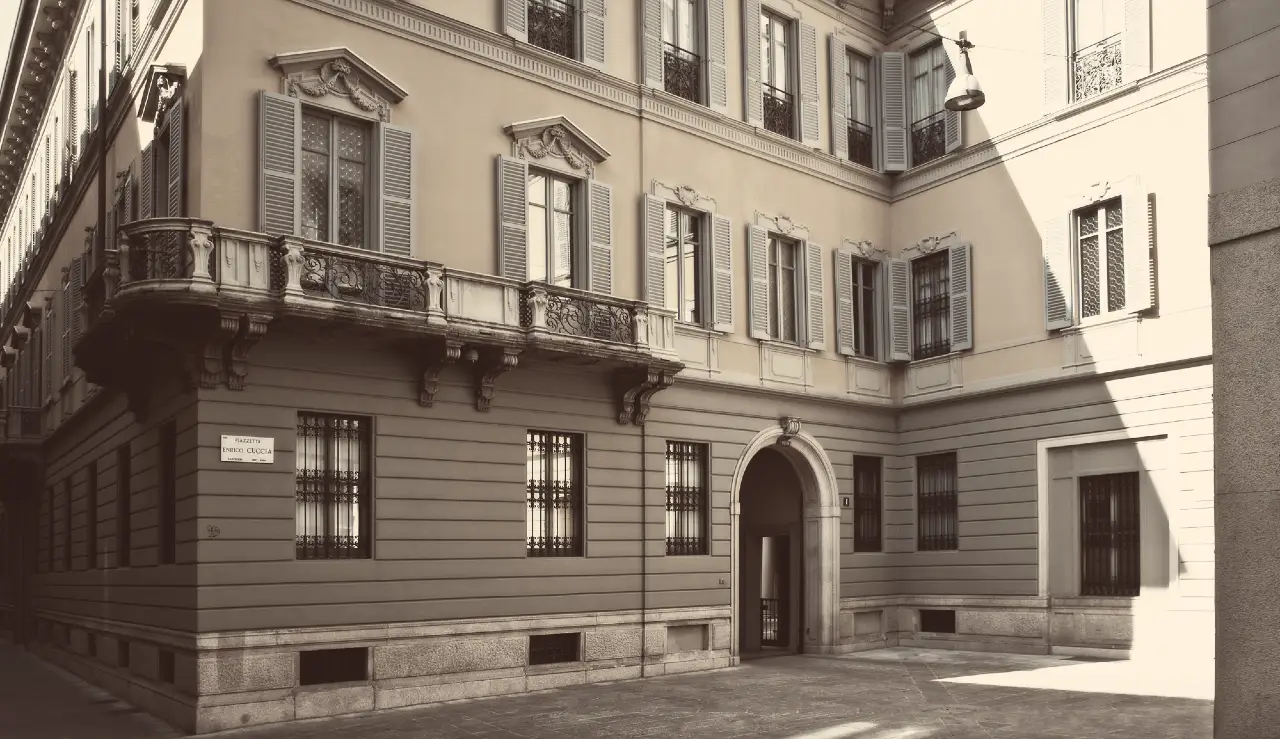 Foto in tonalità seppia di un edificio storico europeo con balconi ornati, finestre a persiana e un ingresso ad arco, situato in un angolo tranquillo e stretto della strada. La luce soffusa del sole proietta ombre sulla facciata.