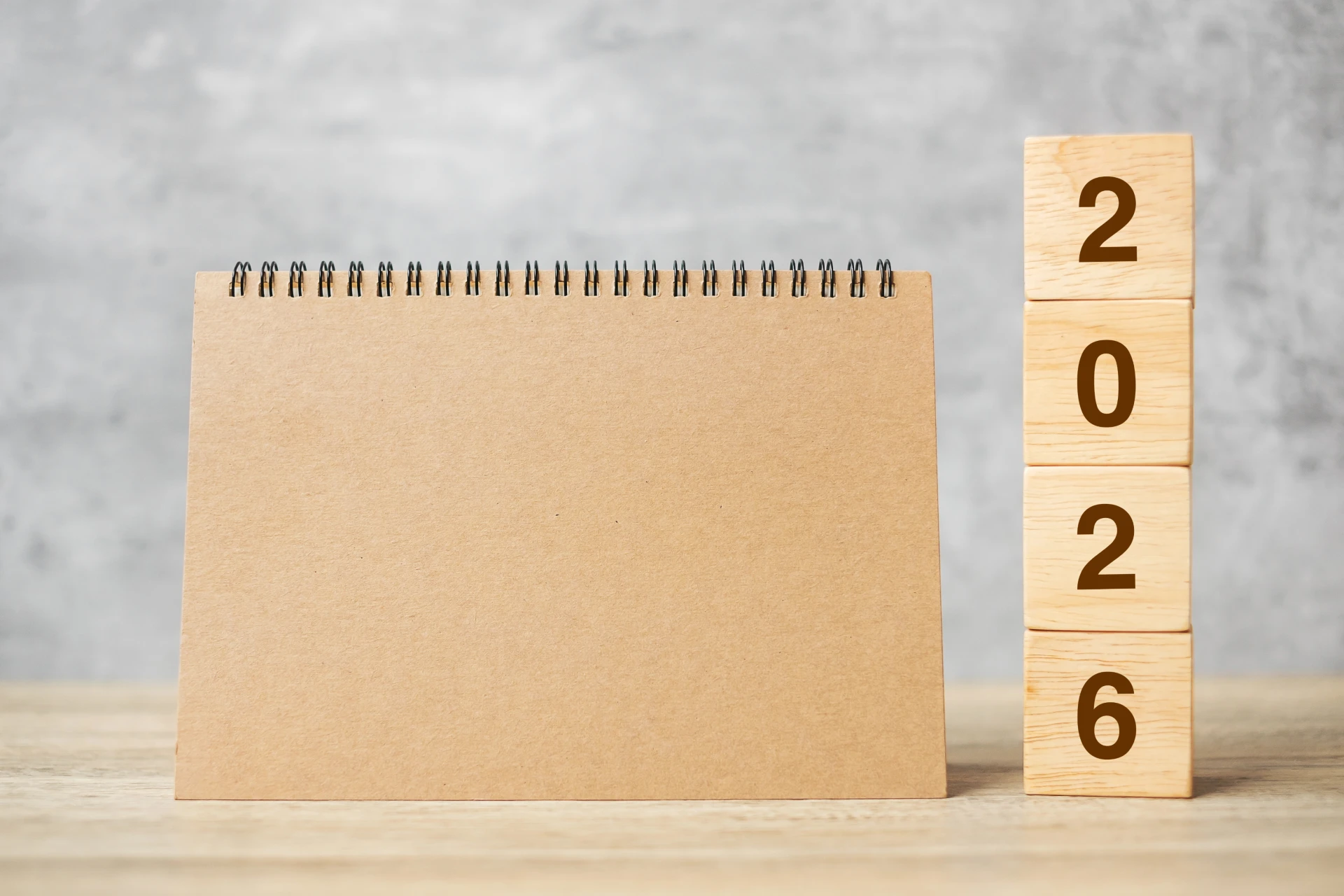 Un calendario da tavolo marrone, rilegato a spirale e vuoto, si trova accanto a una pila verticale di blocchi di legno con la scritta "2026", che simboleggia lo scenario dei mercati del 2026 su una superficie di legno chiaro con uno sfondo grigio.