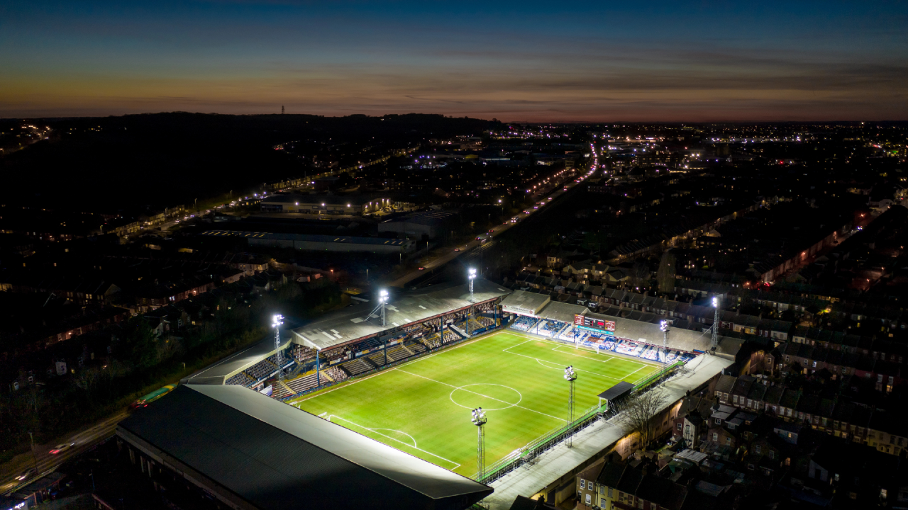 Veduta aerea notturna di uno stadio di calcio vuoto, il campo appare come un rettangolo luminoso immerso nel tessuto urbano circostante.