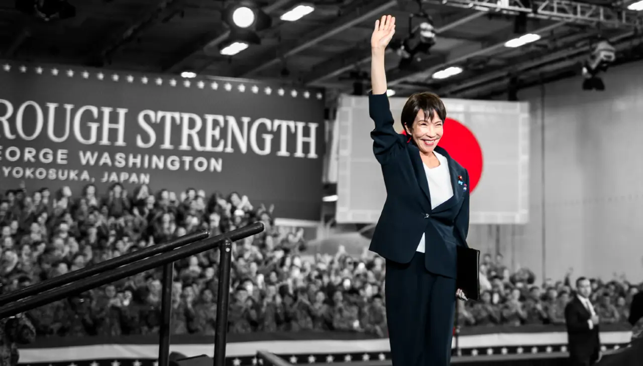 Una donna in abito scuro saluta in piedi su un palco davanti a una folla acclamante, con una grande bandiera giapponese e il testo "ROUGH STRENGTH GEORGE WASHINGTON YOKOSUKA, JAPAN" sullo sfondo.