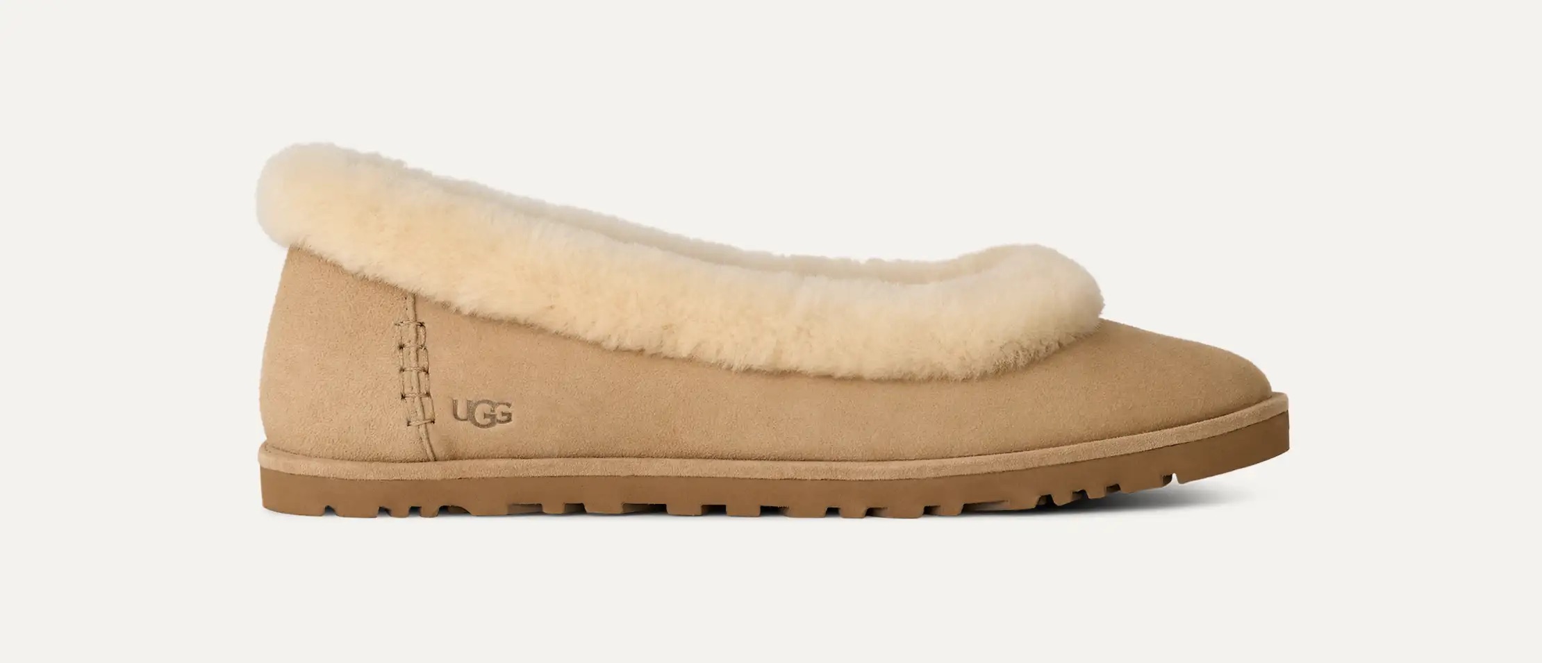 Una pantofola UGG abbronzata con suola in gomma marrone chiaro e morbida fodera in shearling color crema lungo l'apertura, vista di lato su sfondo bianco.