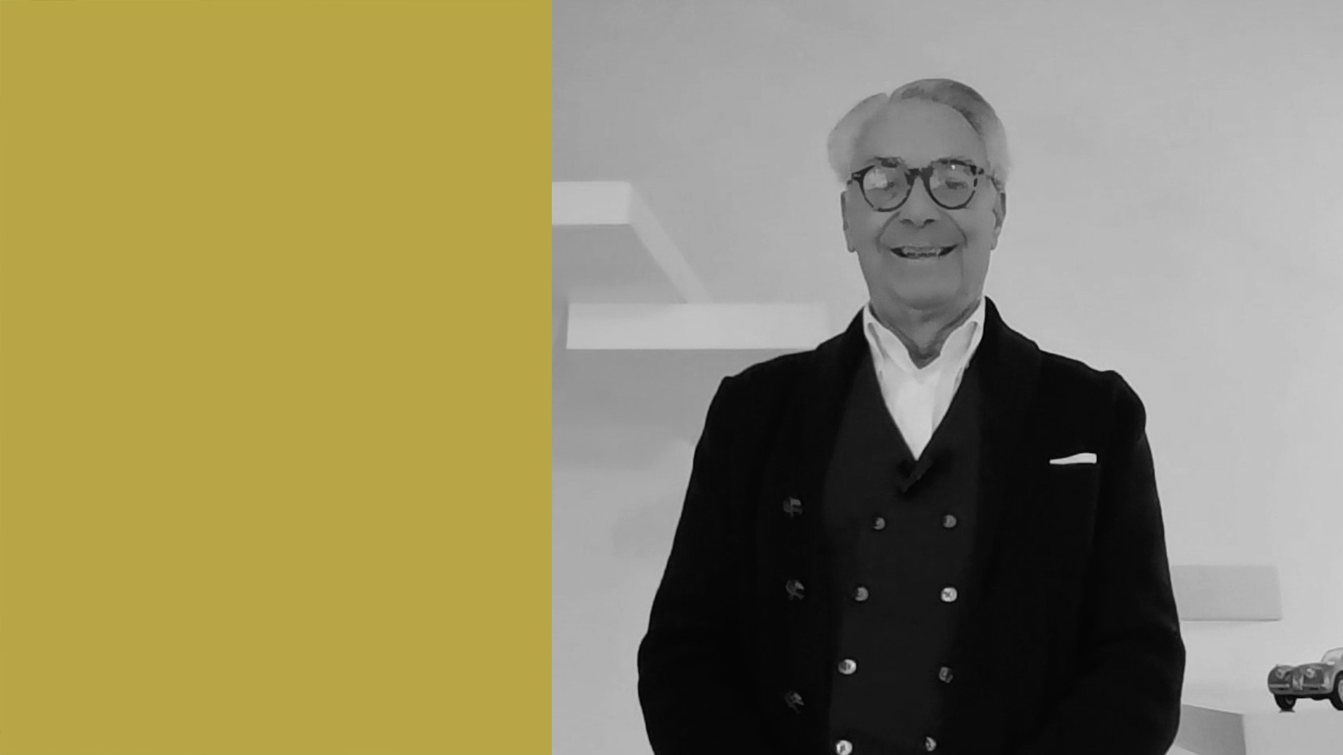 Gianluigi Vignola di ADEMY vestito con un abito scuro e una camicia bianca, si trova in un ambiente interno. Il lato sinistro dell'immagine è caratterizzato da un blocco solido giallo senape, mentre il lato destro mostra uno sfondo bianco minimo.