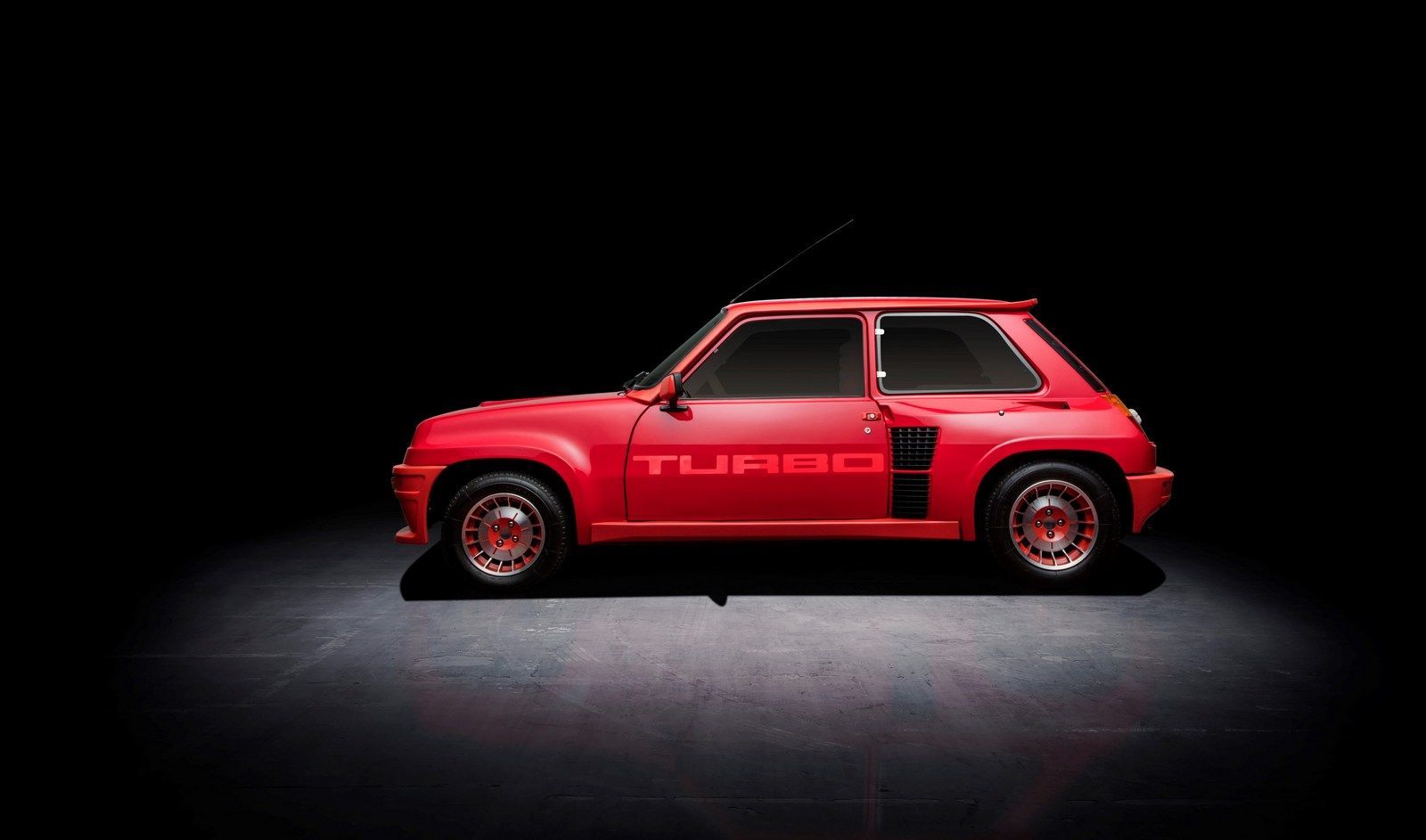 Una classica auto sportiva Renault 5 Turbo rossa è raffigurata in uno studio con sfondo nero, le sue linee audaci e le caratteristiche distintive della Renault 5 Turbo sono messe in risalto dall'illuminazione dall'alto.
