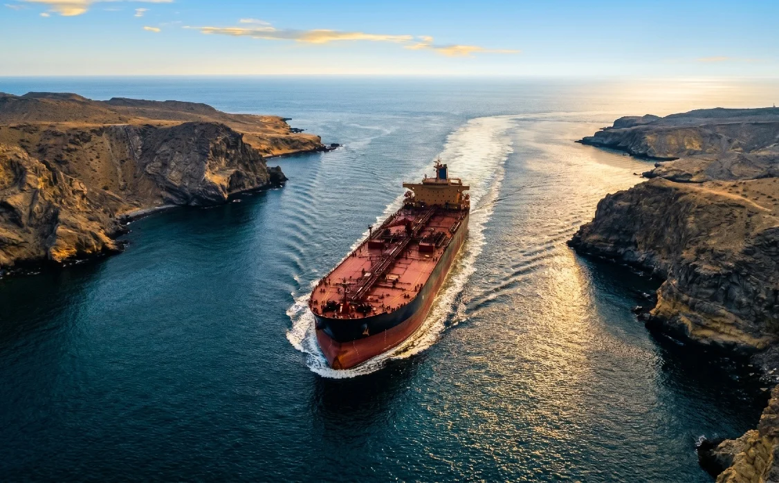 Veduta aerea zenitale di una massiccia nave petroliera rossa e nera che naviga in acque profonde tra alte scogliere rocciose all'alba o al tramonto, lasciando una scia luminosa. L'immagine evoca il passaggio attraverso lo Stretto di Hormuz durante una crisi energetica geopolitica.