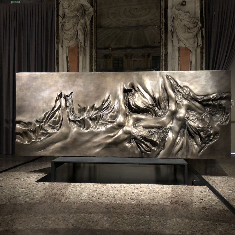 Scultura orizzontale in metallo lucido con figure umane stilizzate, esposta su una panca in un ambiente classico con tendaggi e statue.