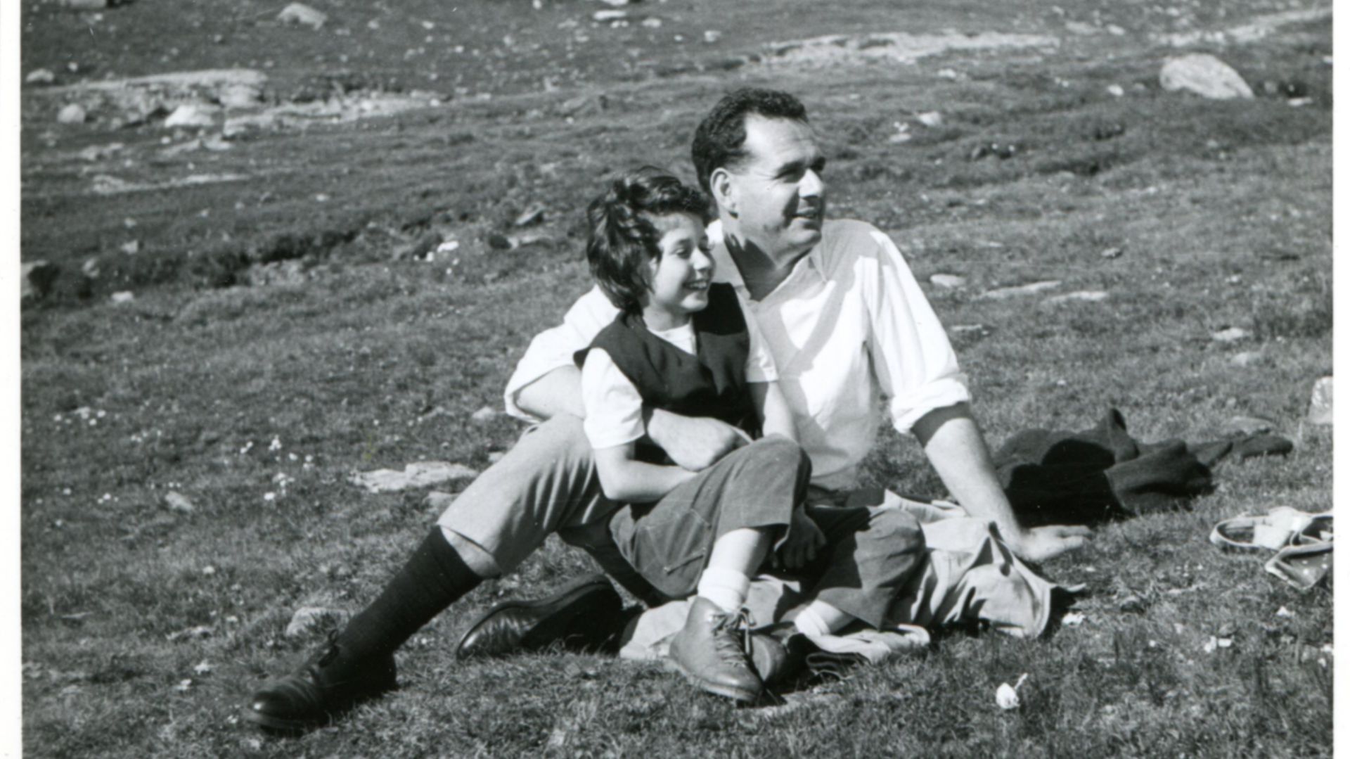 Uomo e bambina seduti sull'erba in montagna, lui la tiene stretta. Foto in bianco e nero.
