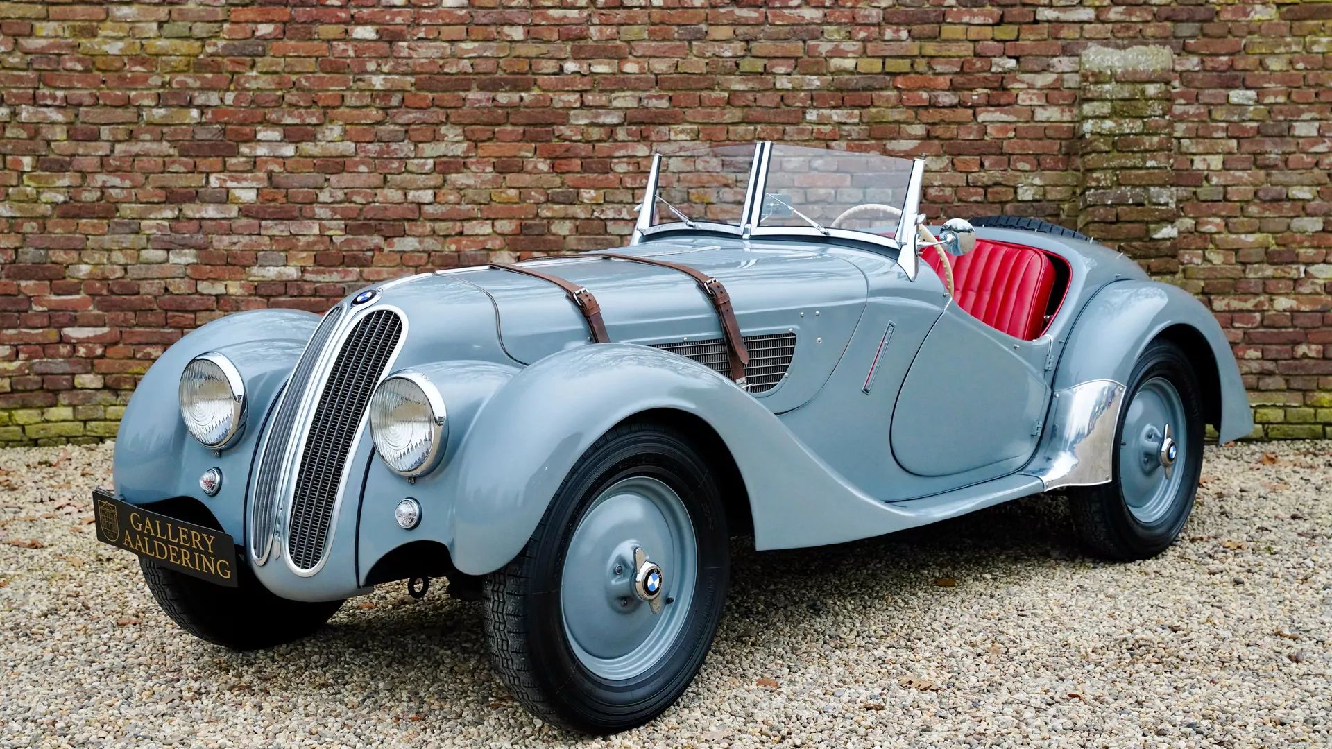 Auto d'epoca BMW grigia cabriolet con interni rossi, cinghie in cuoio sul cofano e muro di mattoni sullo sfondo.