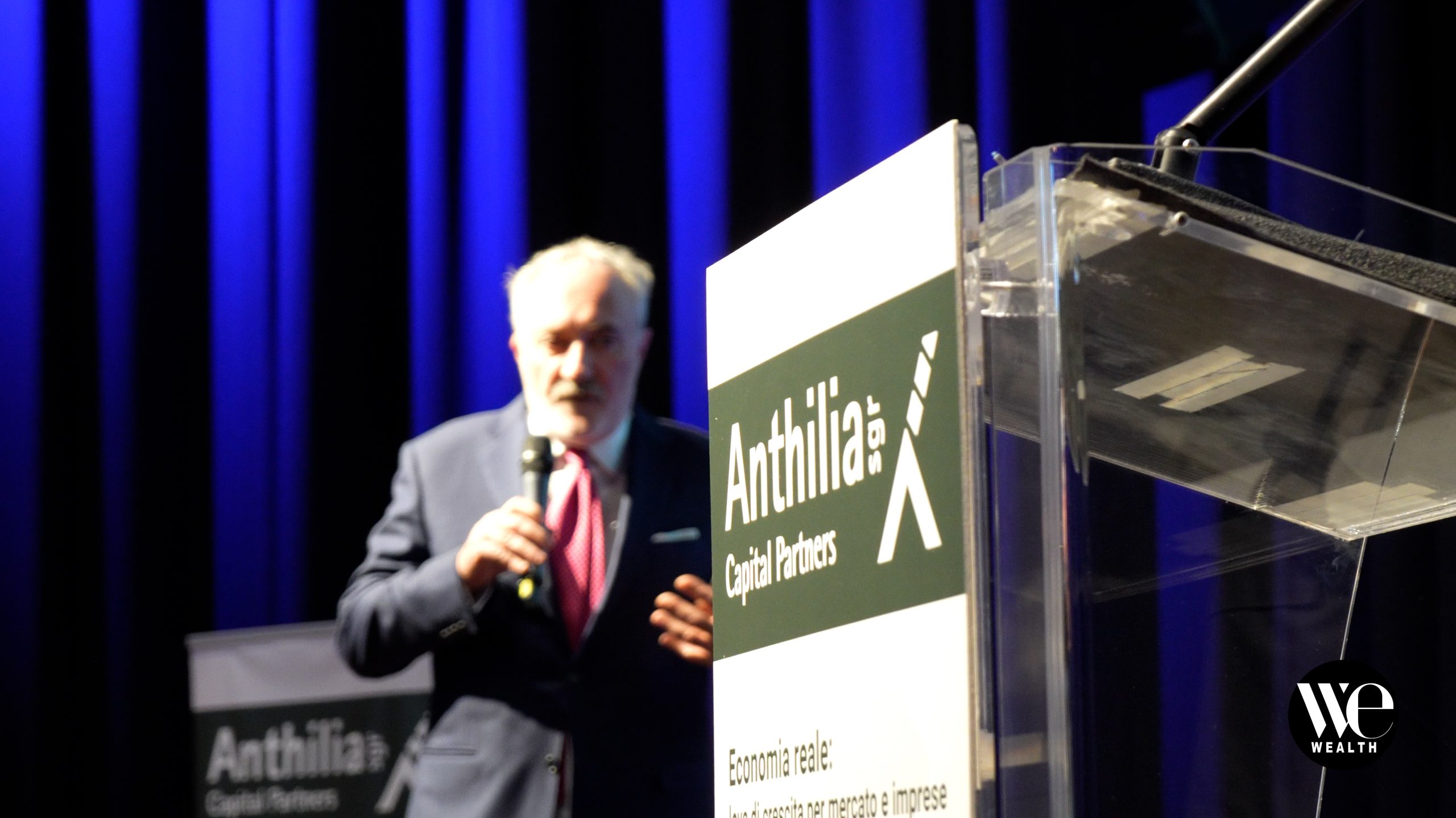 Uomo al microfono accanto a leggio con logo "Anthilia Capital Partners" su sfondo blu.