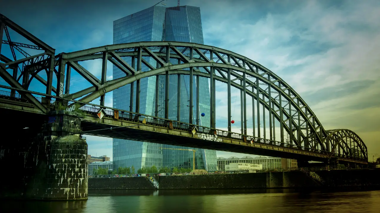 Ponte di ferro ad arco sul fiume, con grattacielo moderno sullo sfondo e cielo nuvoloso.