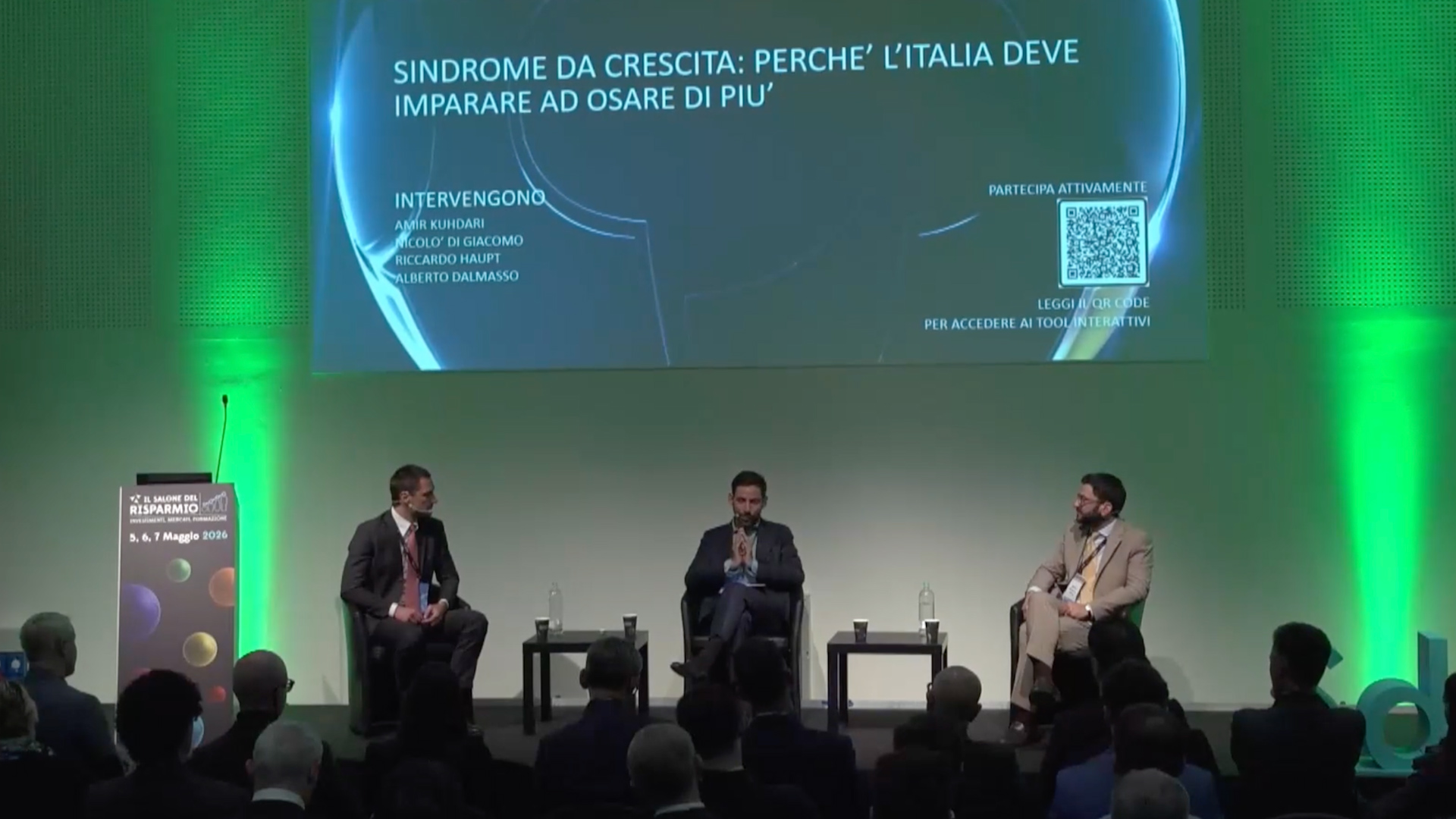 Tre uomini seduti su sedie durante una conferenza con pubblico, schermo con titolo e QR code.