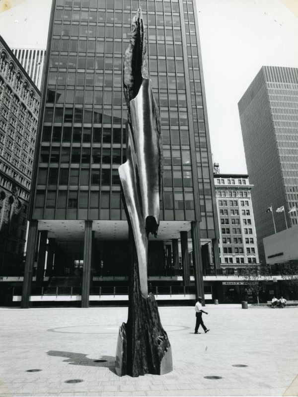 Scultura astratta in metallo lucido su base scura, con grattacieli sullo sfondo e un uomo che cammina.